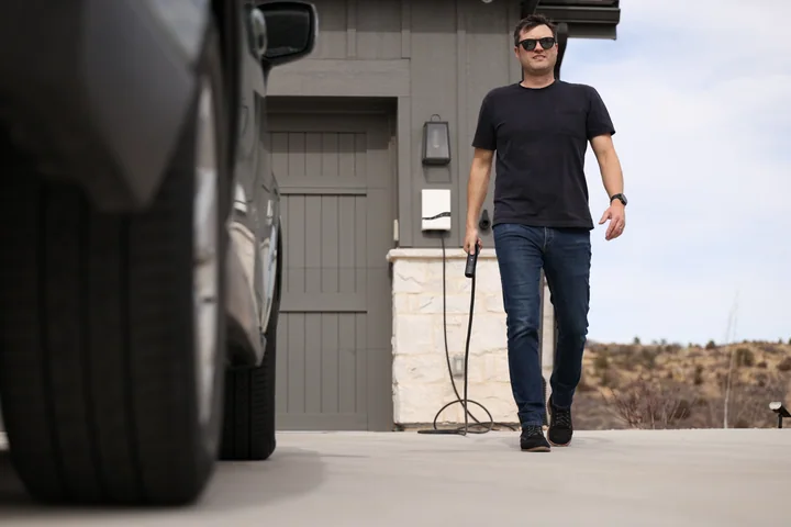 A person carries a charging connector beside a garage with a white Emporia Pro charger.